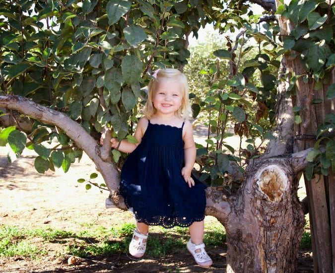 Portrait of pre-school age Aurora Learned, a blond girl in a navy blue sundress, sitting on a tree's lower branch and smiling