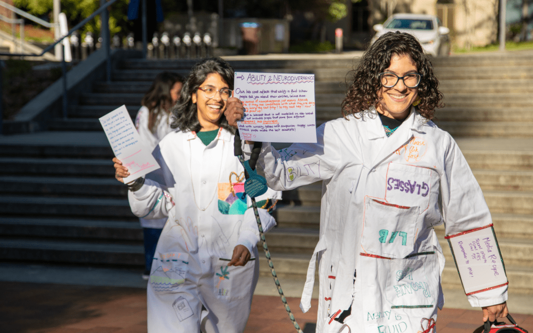 Two students in decorated lab coats hold up signs. The first one is titled "ability & neurodivergance." The second is illegible.